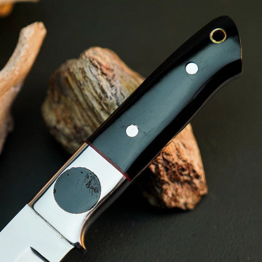A close-up of a knife handle with a glossy black grip, silver bolster, and brass eyelet, resting against natural wood pieces on a dark surface.