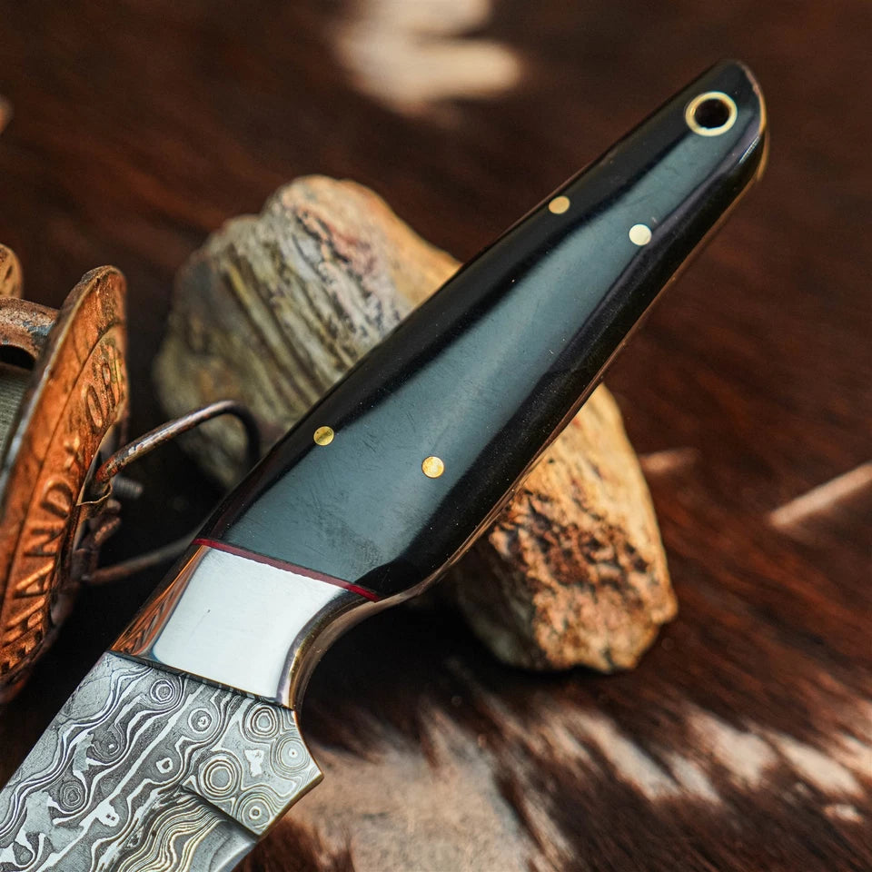 A close-up of a knife with a patterned Damascus steel blade and a dark handle featuring brass pins, resting on rustic stones over a brown textured surface.