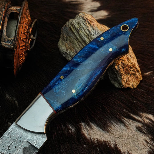 A close-up of a knife with a polished blue marbled handle and patterned steel blade, resting on dark fur with rocks and a copper-colored object in the background.