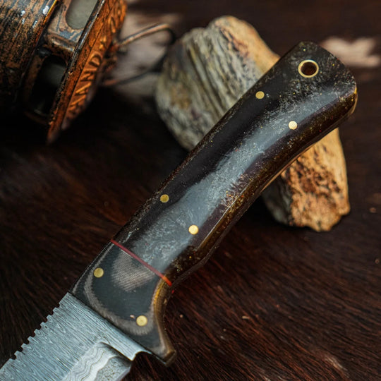 A close-up of a handcrafted knife with a dark brown handle featuring brass pins and a Damascus steel blade, resting on a dark wooden surface with rustic stones.