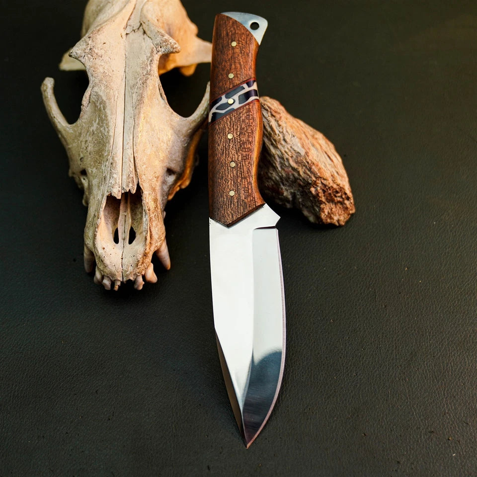 A fixed-blade knife with a polished silver blade and a brown wood-grain handle is displayed vertically on a dark textured surface, styled with a deer skull and a piece of wood behind it.