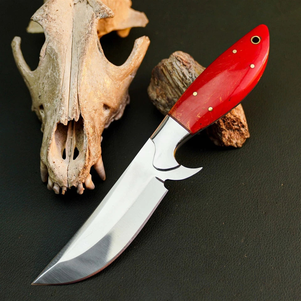 A fixed-blade knife with a polished silver blade and a vibrant red handle, styled on a dark textured surface next to animal skull and wood props.