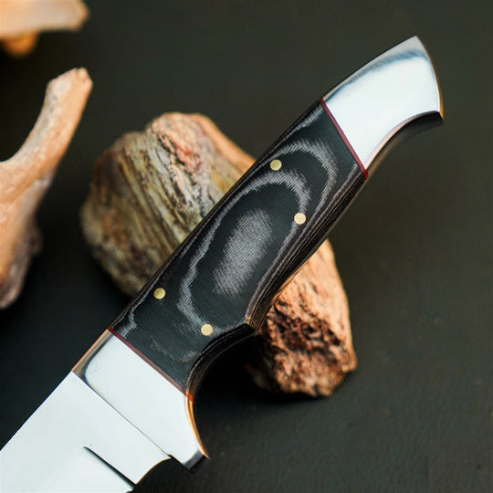A close-up of a fixed-blade knife with a polished silver steel blade and handle, featuring a black textured inlay with a wavy pattern and brass pins, resting on rustic stones against a dark background.