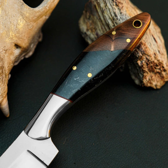 A close-up of a custom knife with a polished silver blade and an inlaid handle made of dark wood, black resin, and gold accents, resting on a dark surface with rustic wood props.