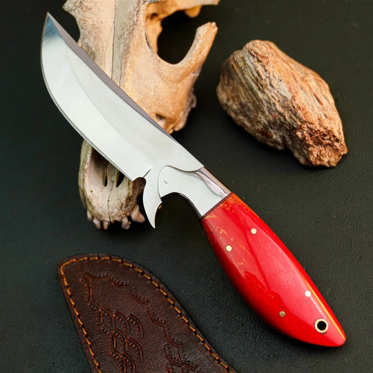 A fixed-blade knife with a polished silver blade and a glossy red handle, displayed diagonally on a dark textured surface with a bone specimen, a rock, and a brown leather sheath.