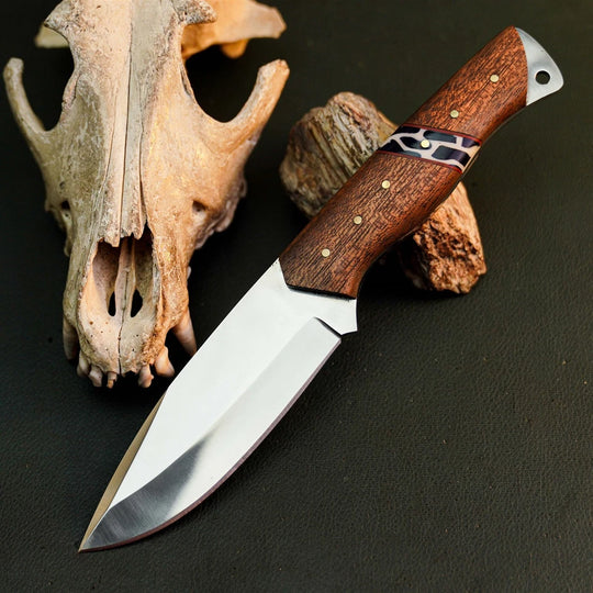 A fixed-blade knife with a polished silver blade and a brown wooden handle with a black and white patterned inlay, displayed on a dark textured surface next to an animal skull and wood pieces.