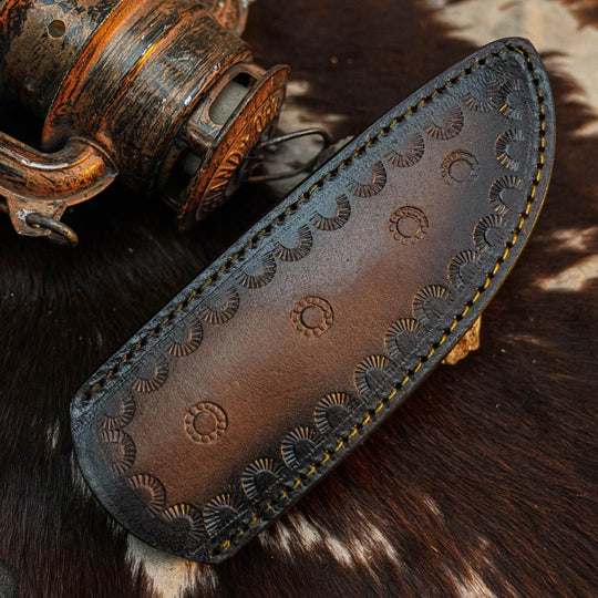 A dark brown leather knife sheath with hand-tooled circular patterns and yellow stitching, displayed on a cowhide surface next to an antique wooden object.