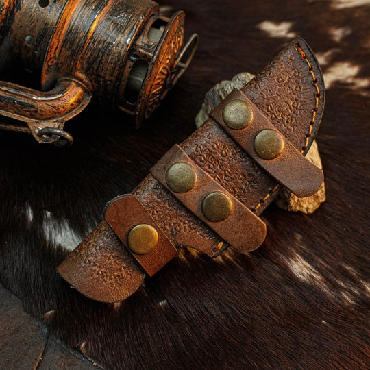 A close-up of a brown leather knife sheath with embossed floral detailing and brass snap closures, placed on a dark brown and white cowhide surface next to a rustic metal object.