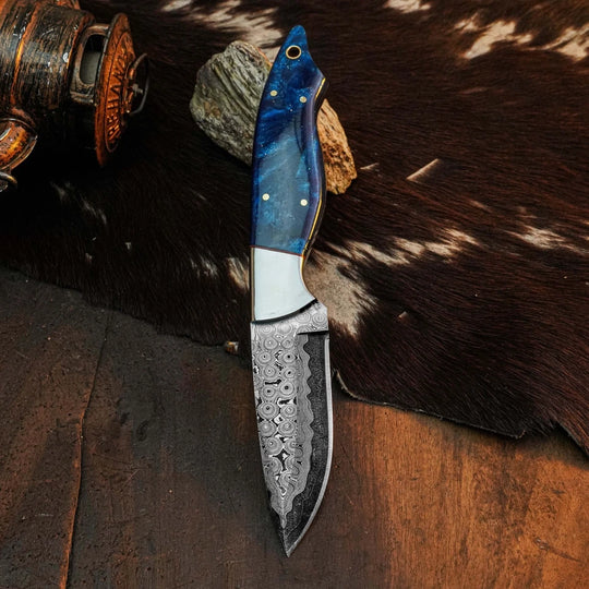 A fixed-blade knife with a patterned steel blade and a blue marbled handle, displayed on a rustic brown surface with a cowhide rug and a wooden object in the background.