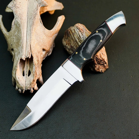 A fixed-blade knife with a polished silver blade and a black handle with brass pins, displayed diagonally on a dark textured surface next to a deer skull and a piece of driftwood.