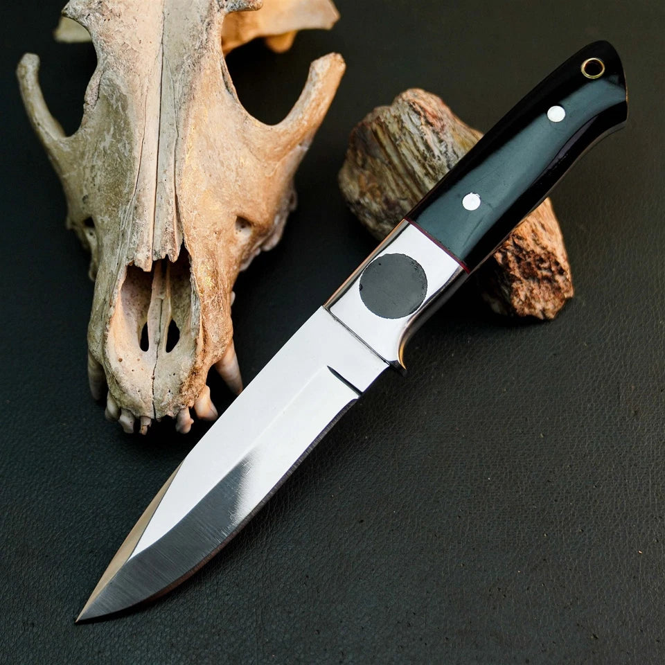 A fixed-blade knife with a polished silver blade and a black handle with silver rivets, displayed diagonally on a dark textured surface next to a bone skull and a piece of wood.