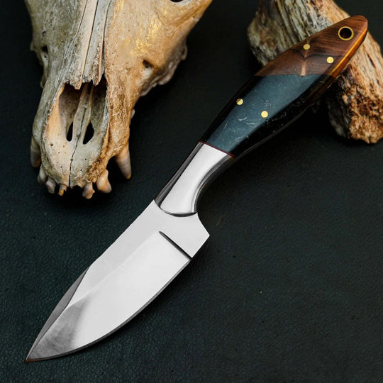 A fixed-blade knife with a satin-finished silver blade and a dark wood handle with a blue-green inlay, displayed diagonally on a dark textured surface next to a weathered animal skull.