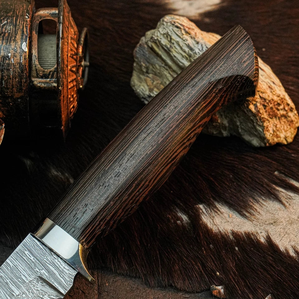 A close-up of a knife with a dark wood handle and patterned steel blade, resting on a brown fur surface with rocks and part of a rustic metal container in the background.