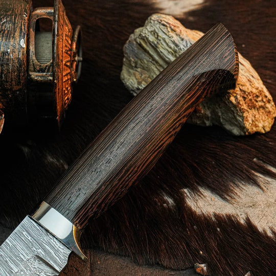 A close-up of a knife with a dark wood handle and patterned steel blade, resting on a brown fur surface with rocks and part of a rustic metal container in the background.