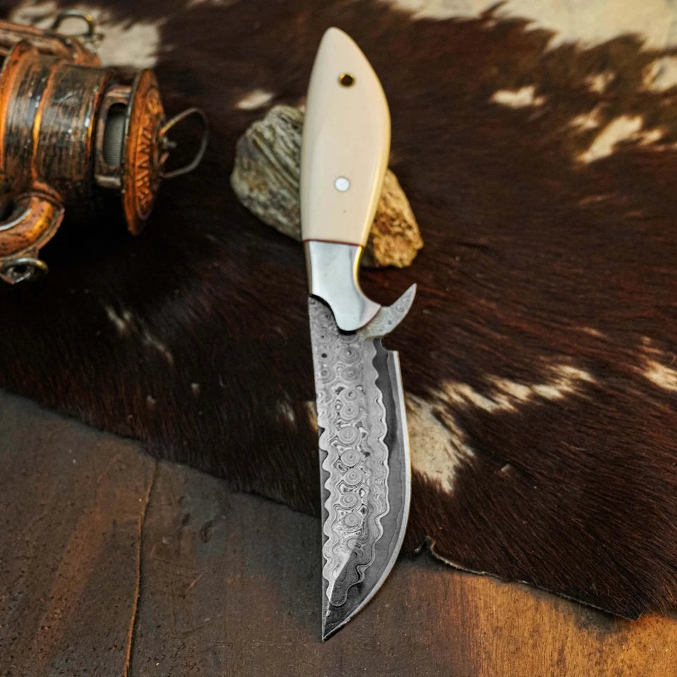 A custom fixed-blade knife with a patterned Damascus steel blade and a cream-colored handle, displayed on a brown and white cowhide rug next to a rustic lantern.