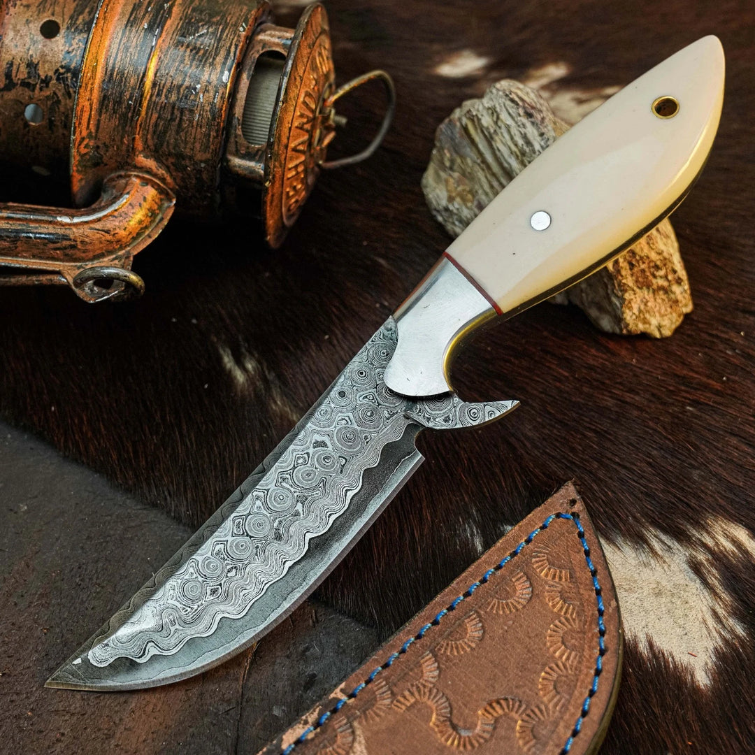 A custom handmade fixed-blade knife with a patterned Damascus steel blade, a silver bolster, and a cream-colored bone handle, displayed on a brown cowhide background next to a leather sheath and a rustic fishing reel.