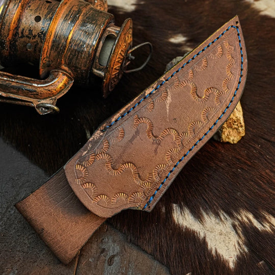 A handcrafted brown leather knife sheath with embossed wave patterns and blue stitching, displayed on a cowhide surface next to a rustic metal object.