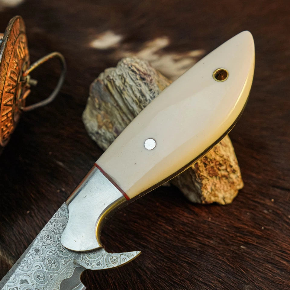 A close-up of a folding pocket knife with a patterned Damascus steel blade and a cream-colored bone handle, resting on a textured rock over a dark wood surface.