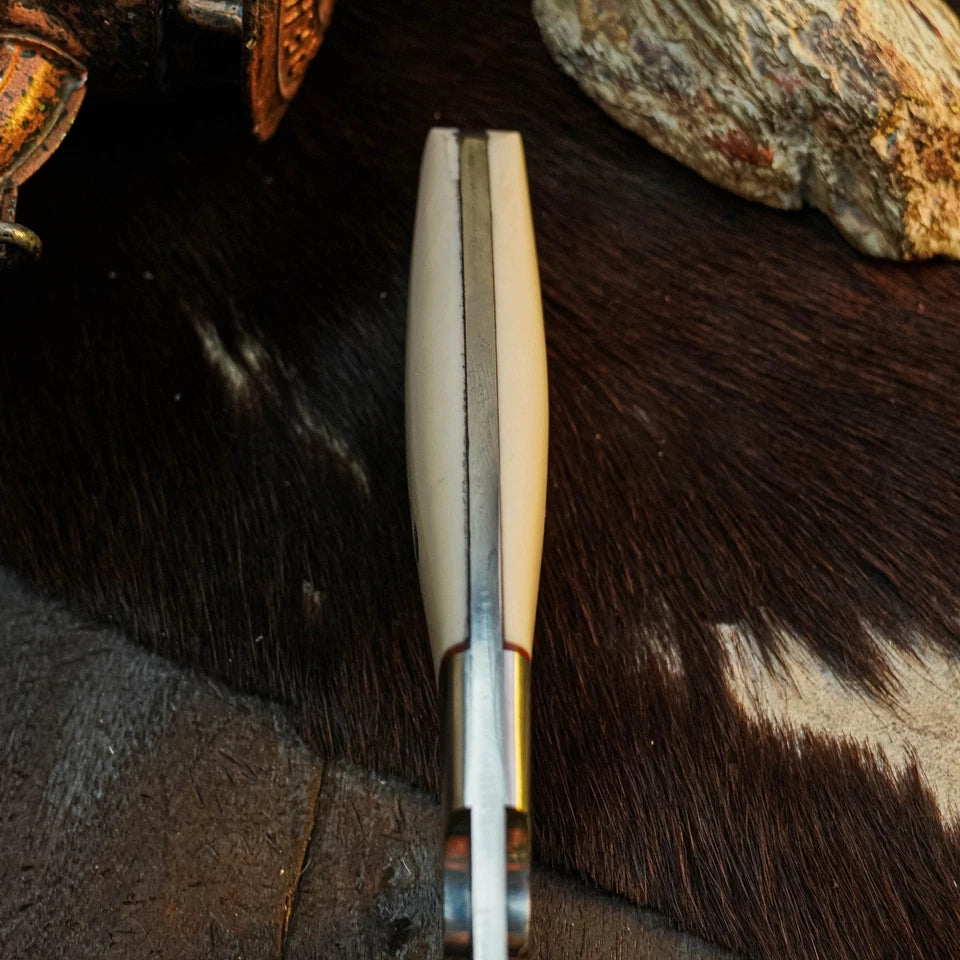 A close-up of a folding pocket knife with a cream-colored bone handle and a silver steel blade, resting on a dark brown and black cowhide surface with rustic stones in the background.
