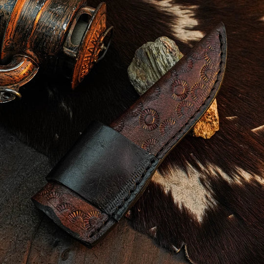 A dark brown leather knife sheath with hand-tooled circular patterns, shown on a rustic background with cowhide and wooden objects.