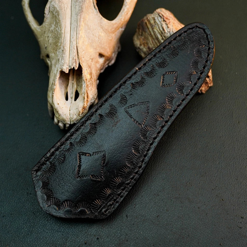 A black leather knife sheath with hand-stitched geometric patterns and scalloped edges, displayed diagonally on a dark textured surface with a deer skull and wood prop in the background.