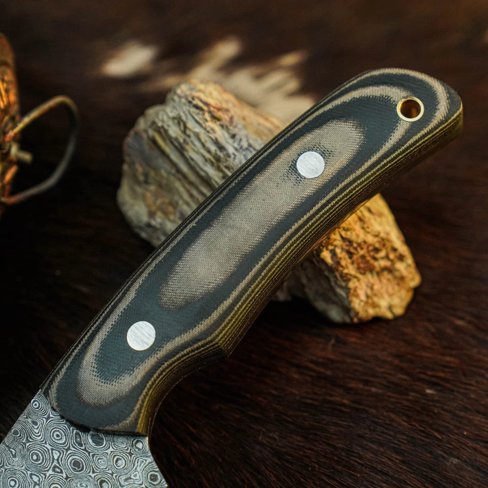 A close-up of a knife handle with a dark gray and black striped pattern and a patterned steel blade, resting on a textured rock against a dark wooden background.