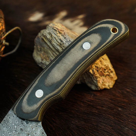 A close-up of a knife handle with a dark gray and black striped pattern and a patterned steel blade, resting on a textured rock against a dark wooden background.