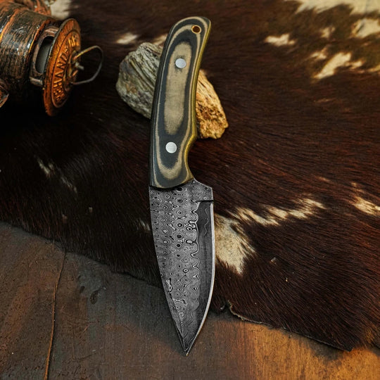 A fixed-blade knife with a patterned steel blade and a dark wood handle with silver rivets, displayed on a brown and white cowhide rug next to a rustic copper object.