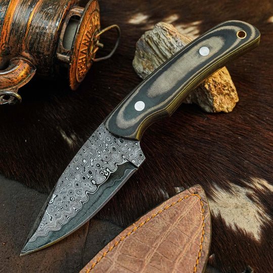 A fixed-blade knife with a patterned Damascus-style steel blade and a contoured handle made of layered black and gray wood, displayed on a brown and white cowhide surface next to a leather sheath.