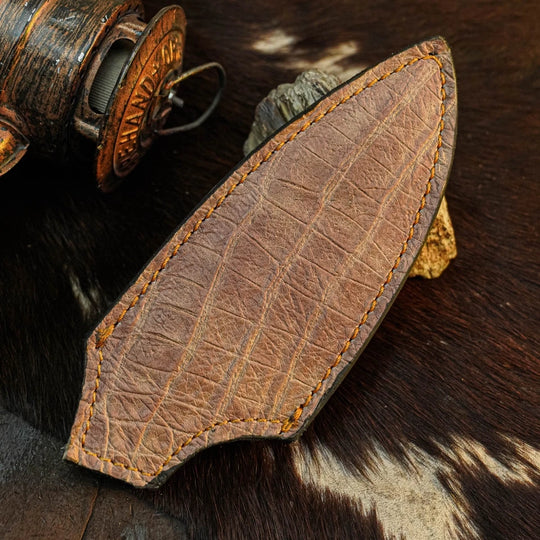 A brown leather knife sheath with a crocodile-embossed texture and contrast stitching, displayed on a dark brown and white cowhide background next to a rustic wooden object.