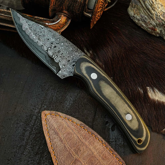 A fixed-blade knife with a patterned Damascus-style steel blade and a dark marbled handle, displayed diagonally on a dark textured surface with a brown leather sheath, fur, and rustic objects in the background.