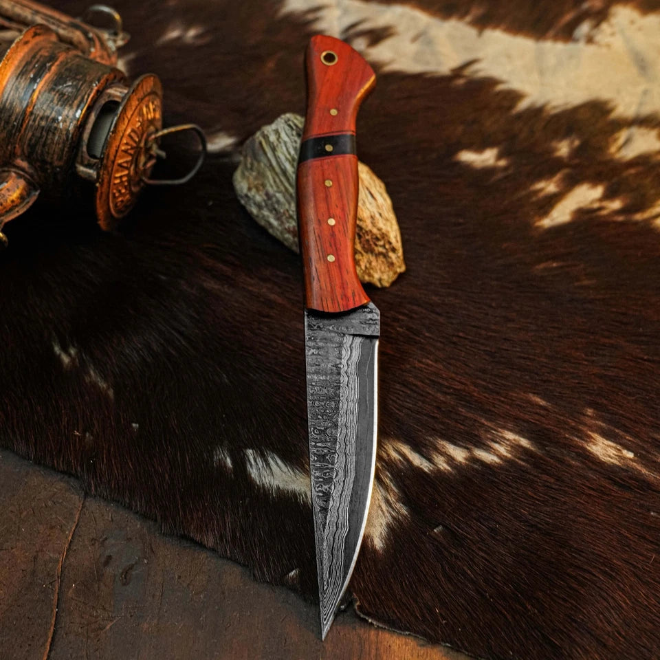 A fixed-blade knife with a patterned steel blade and a reddish-brown wooden handle, displayed on a brown and white cowhide rug next to a rustic lantern.