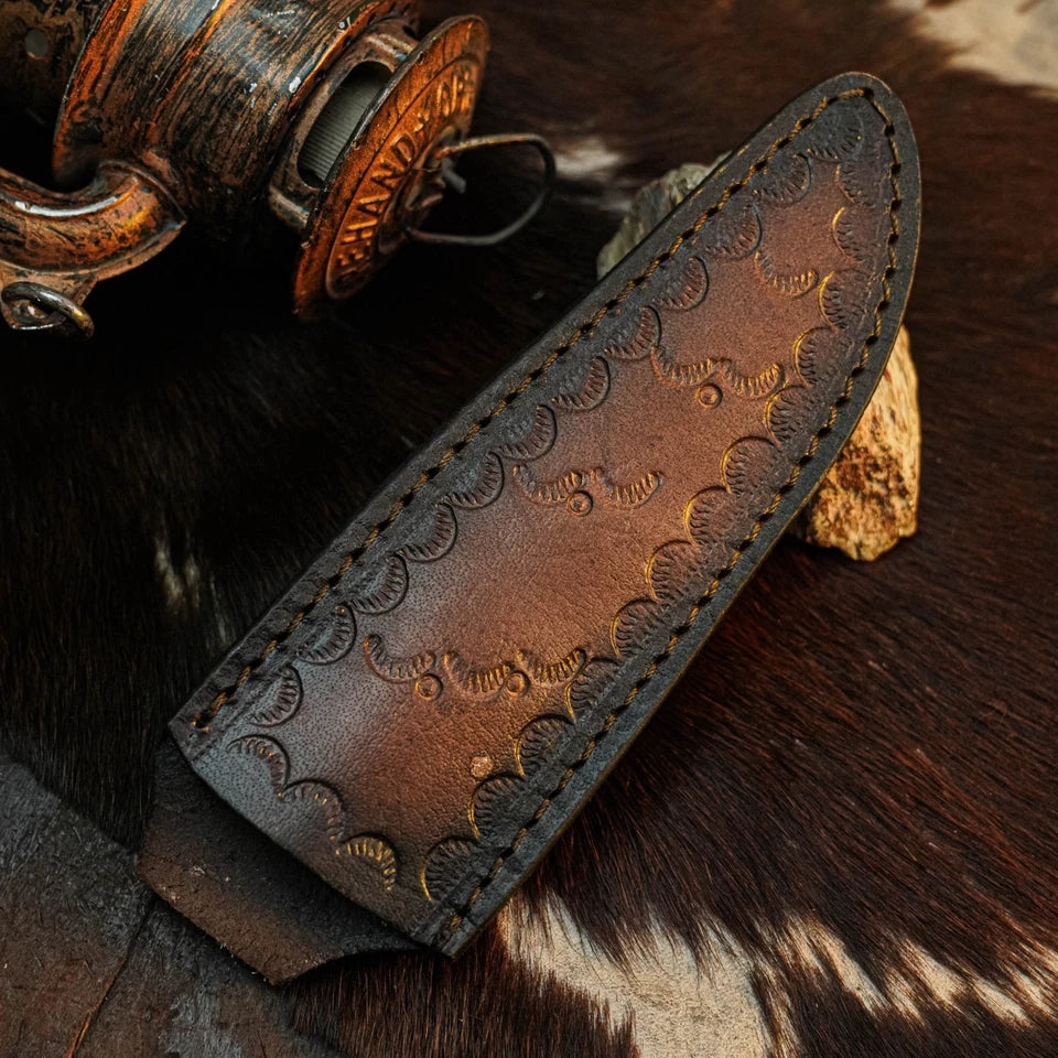 A close-up of a brown leather knife sheath with embossed decorative patterning and visible stitching, placed on a dark brown and white animal hide with rustic metal props in the background.