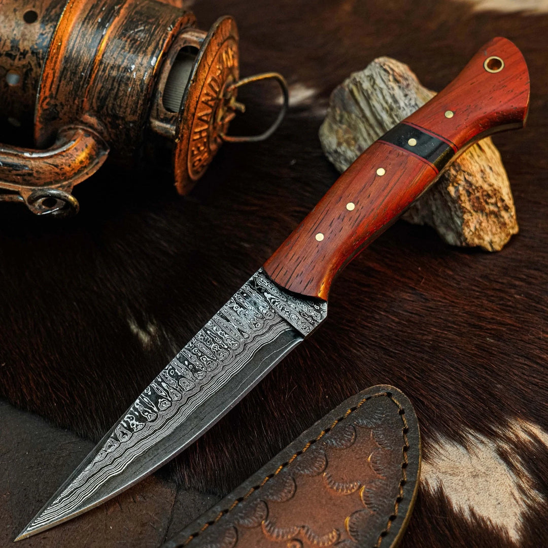 A custom Damascus steel fixed-blade knife with a reddish-brown wooden handle and brass pins, displayed diagonally on a brown and white cowhide rug next to a leather sheath and a vintage lantern.
