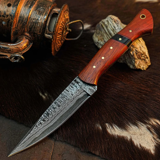 A fixed-blade knife with a patterned Damascus steel blade and a reddish-brown wooden handle, displayed diagonally on a dark brown fur surface with a rustic lantern prop in the background.