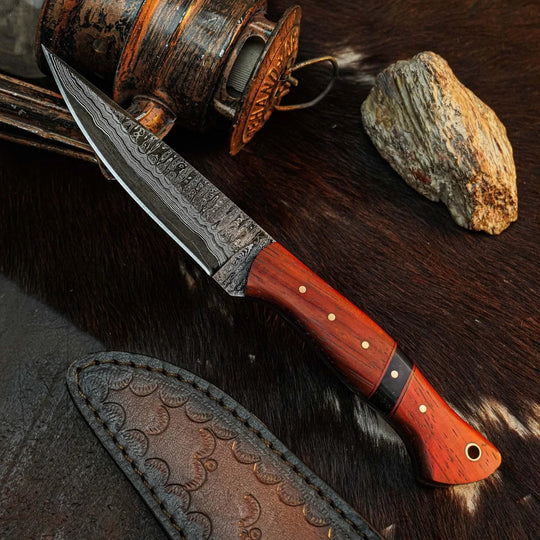 A fixed-blade knife with a patterned steel blade and a reddish-brown wooden handle with brass pins, displayed diagonally on dark animal fur alongside a leather sheath, a rustic wooden lantern, and a rock.