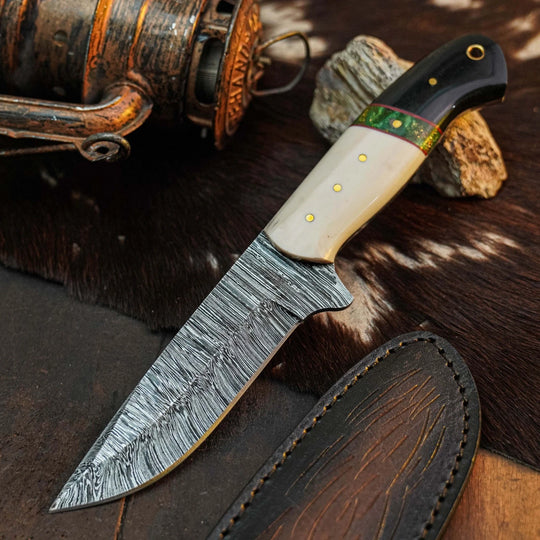 A fixed-blade knife with a patterned steel blade, a cream-colored bone handle, and a black handle with brass pins and a green inlay, displayed next to its dark brown leather sheath on a rustic background with a fishing reel and cowhide.