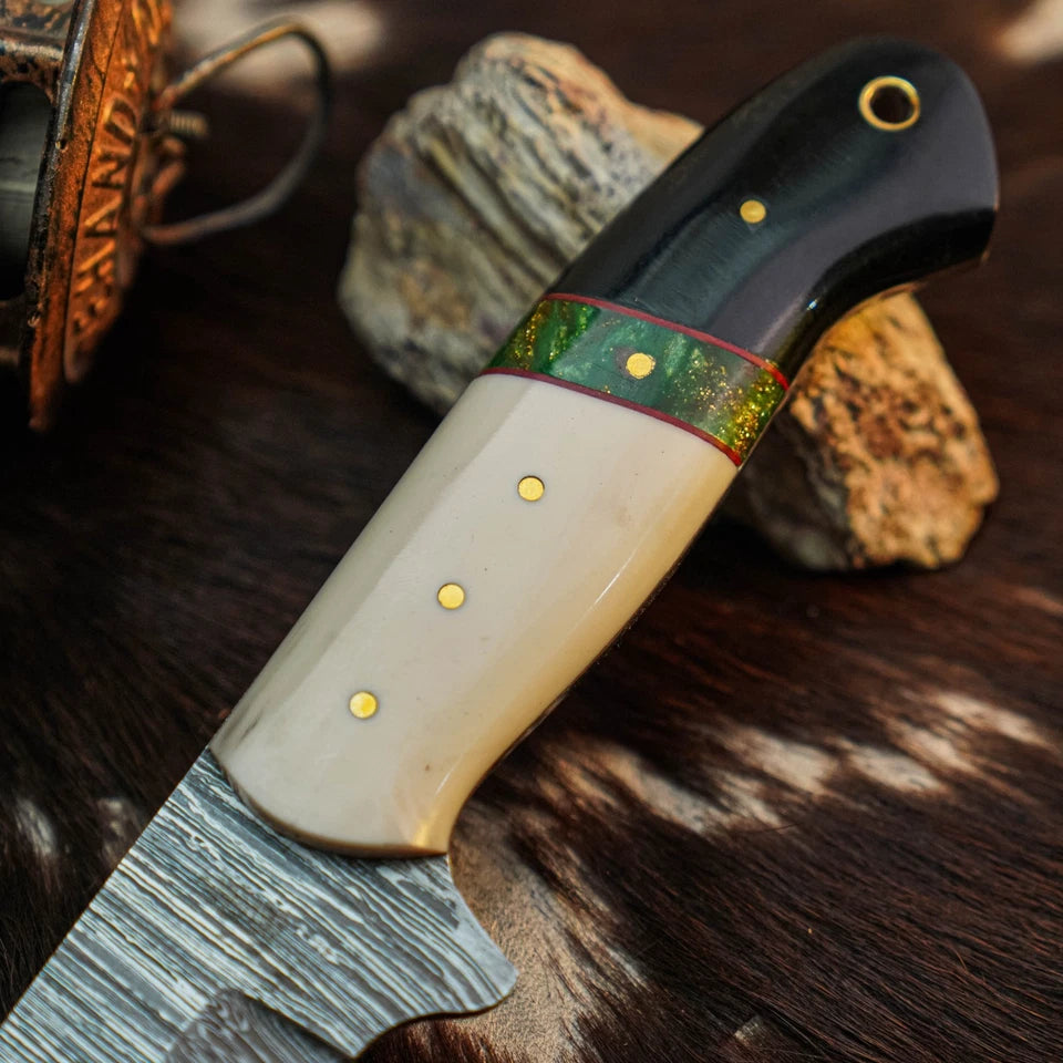 A close-up of a handcrafted knife with a textured steel blade, a cream-colored bone handle, a black end cap, and a green patterned inlay, resting on a dark fur surface with rocks in the background.