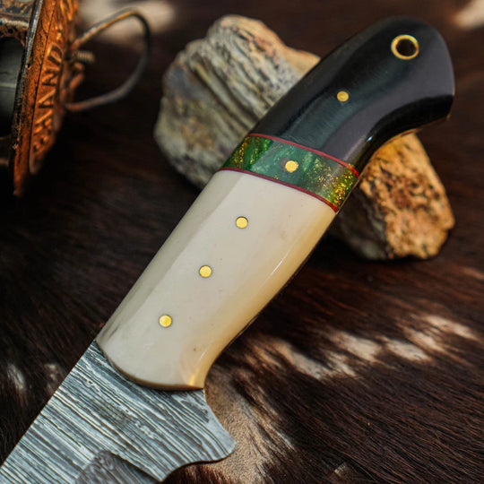 A close-up of a handcrafted knife with a textured steel blade, a cream-colored bone handle, a black end cap, and a green patterned inlay, resting on a dark fur surface with rocks in the background.