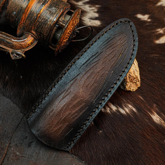 A dark brown leather knife sheath with visible stitching and engraved texture, displayed on a cowhide surface next to a rustic metal object.