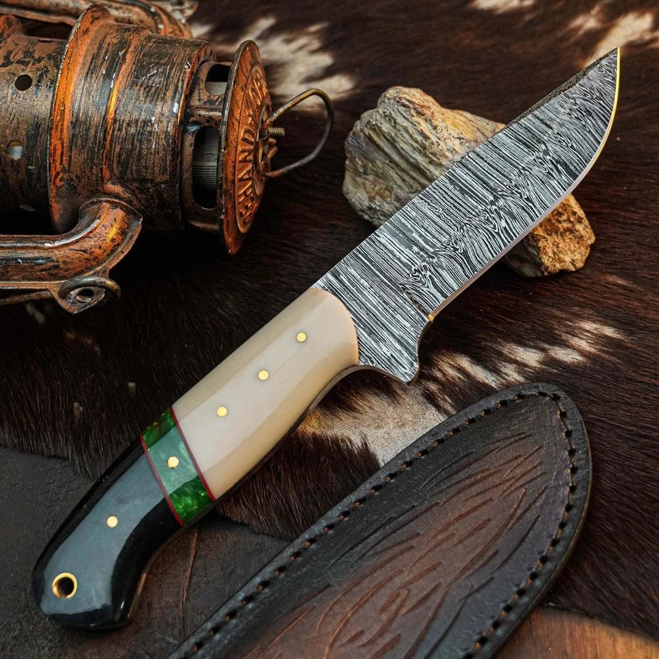 A fixed-blade knife with a patterned steel blade, cream-colored handle scales, a black end cap, and a green accent ring, displayed diagonally on a brown and black cowhide rug next to its dark brown leather sheath. A rustic fishing reel and a small rock are styled as props in the background.