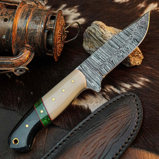 A fixed-blade knife with a patterned steel blade, cream-colored handle scales, a black end cap, and a green accent ring, displayed diagonally on a brown and black cowhide rug next to its dark brown leather sheath. A rustic fishing reel and a small rock are styled as props in the background.