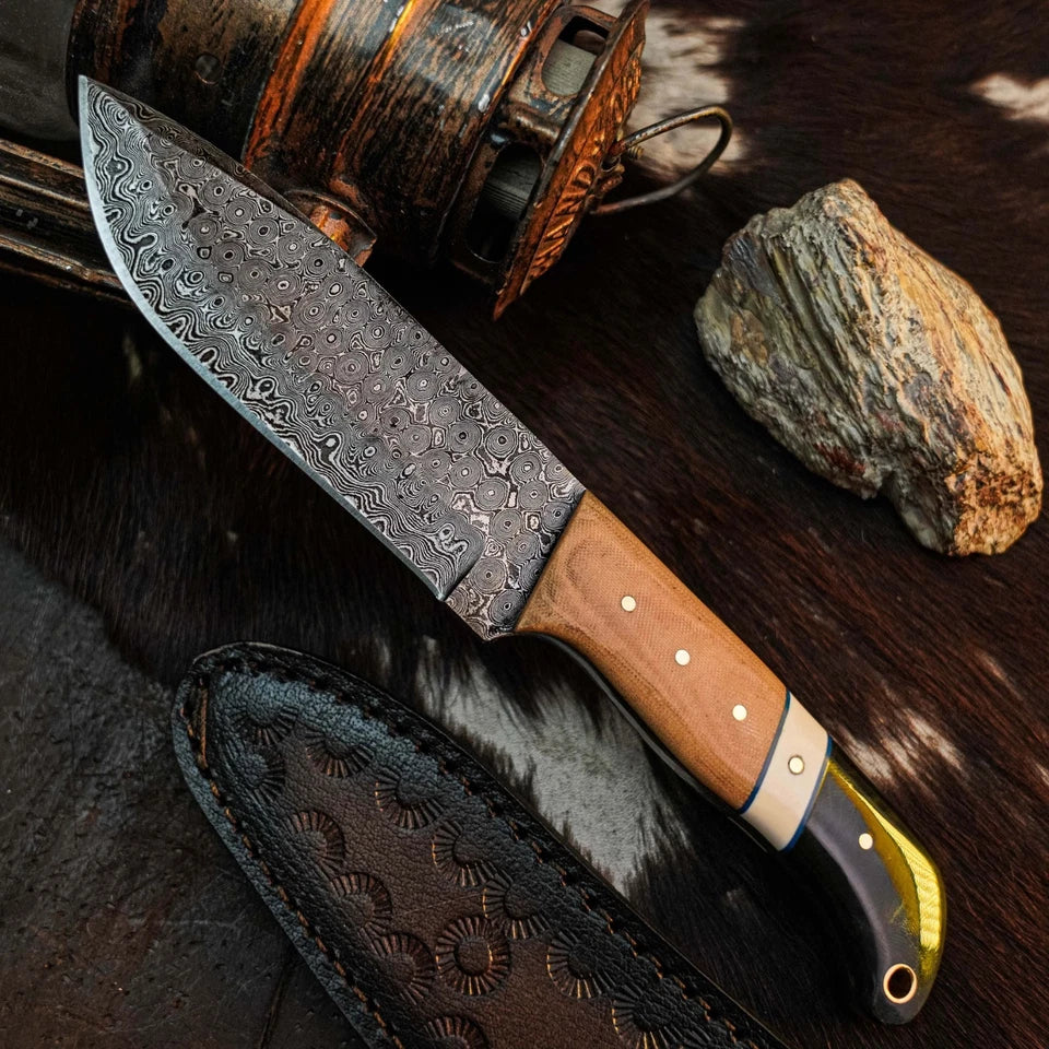 A fixed-blade knife with a patterned Damascus-style steel blade, a light brown wood handle, and a black-and-gold bolster, displayed diagonally on a dark textured surface with a leather sheath, rustic bottle, and stone nearby.