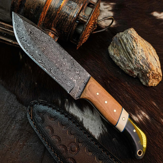 A fixed-blade knife with a patterned Damascus-style steel blade, a light brown wood handle, and a black-and-gold bolster, displayed diagonally on a dark textured surface with a leather sheath, rustic bottle, and stone nearby.