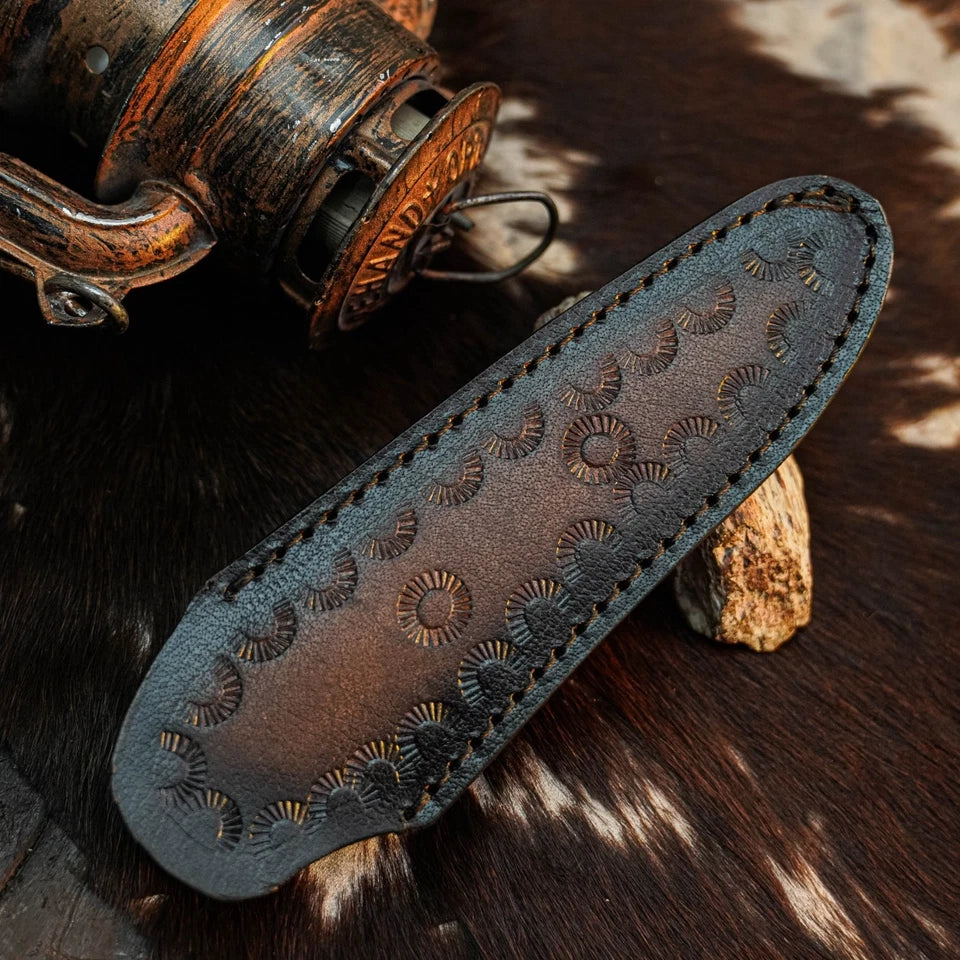 A dark brown leather knife sheath with hand-stitched edges and embossed circular sunburst patterns, displayed on a cowhide surface next to a rustic wooden object.