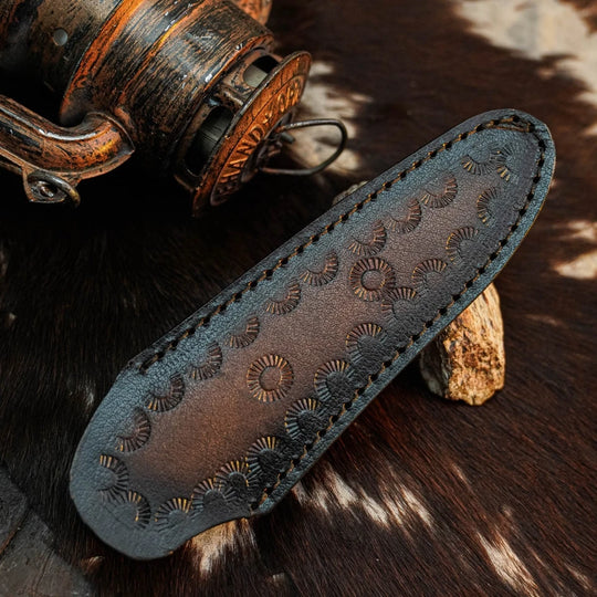 A dark brown leather knife sheath with hand-stitched edges and embossed circular sunburst patterns, displayed on a cowhide surface next to a rustic wooden object.