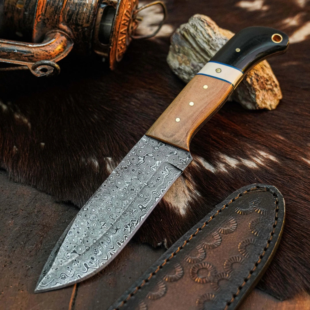 A fixed-blade knife with a patterned steel blade, a wooden handle, and a black handle end, displayed on a brown cowhide surface with a leather sheath and rustic props.