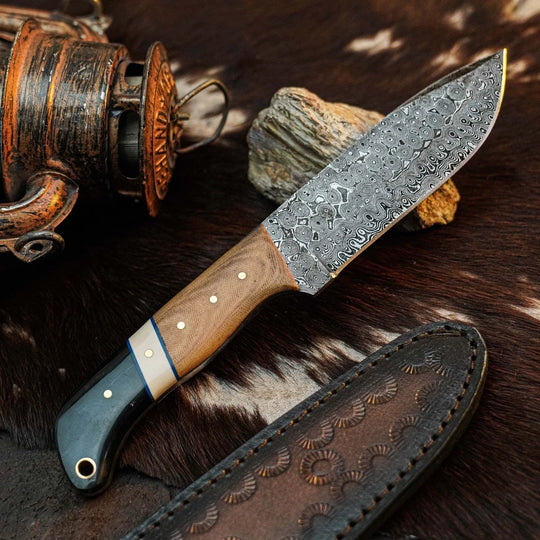 A fixed-blade knife with a patterned Damascus steel blade, a light brown wood handle, and a dark green inlay, displayed diagonally on a brown and white cowhide surface next to its dark brown leather sheath.