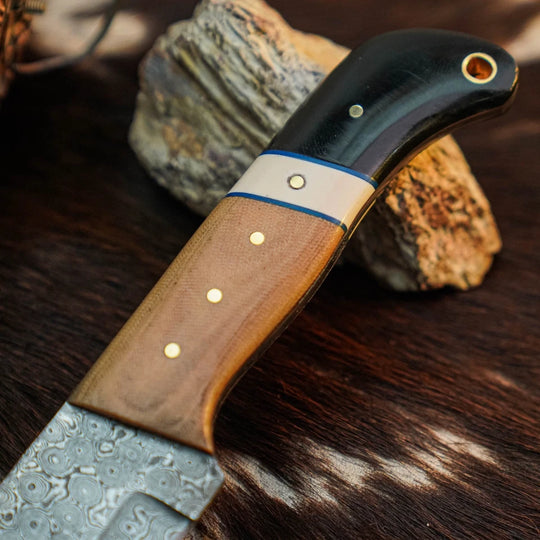 A close-up of a handcrafted knife with a Damascus steel blade, a brown wood handle, and a black horn pommel, resting on a dark textured surface with rocks in the background.