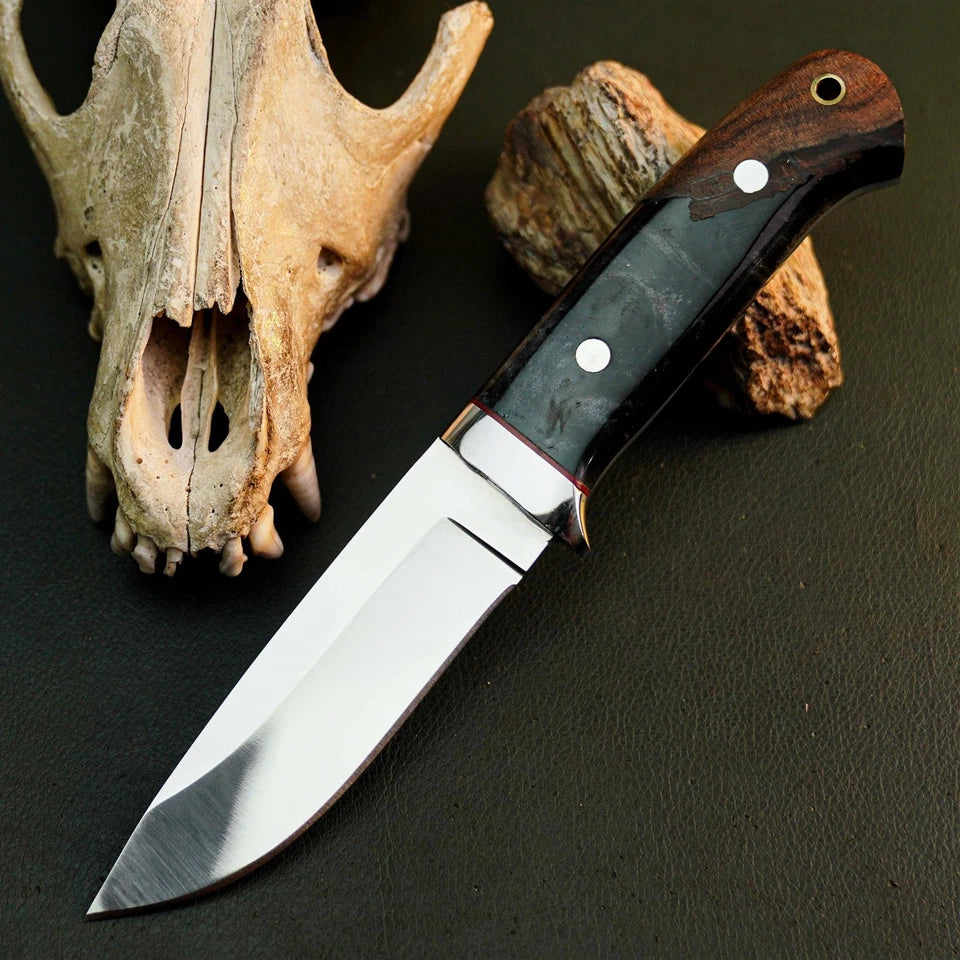 A fixed-blade knife with a polished silver blade and a dark wood handle with a black inlay, displayed diagonally on a dark textured surface next to animal skull and bone props.